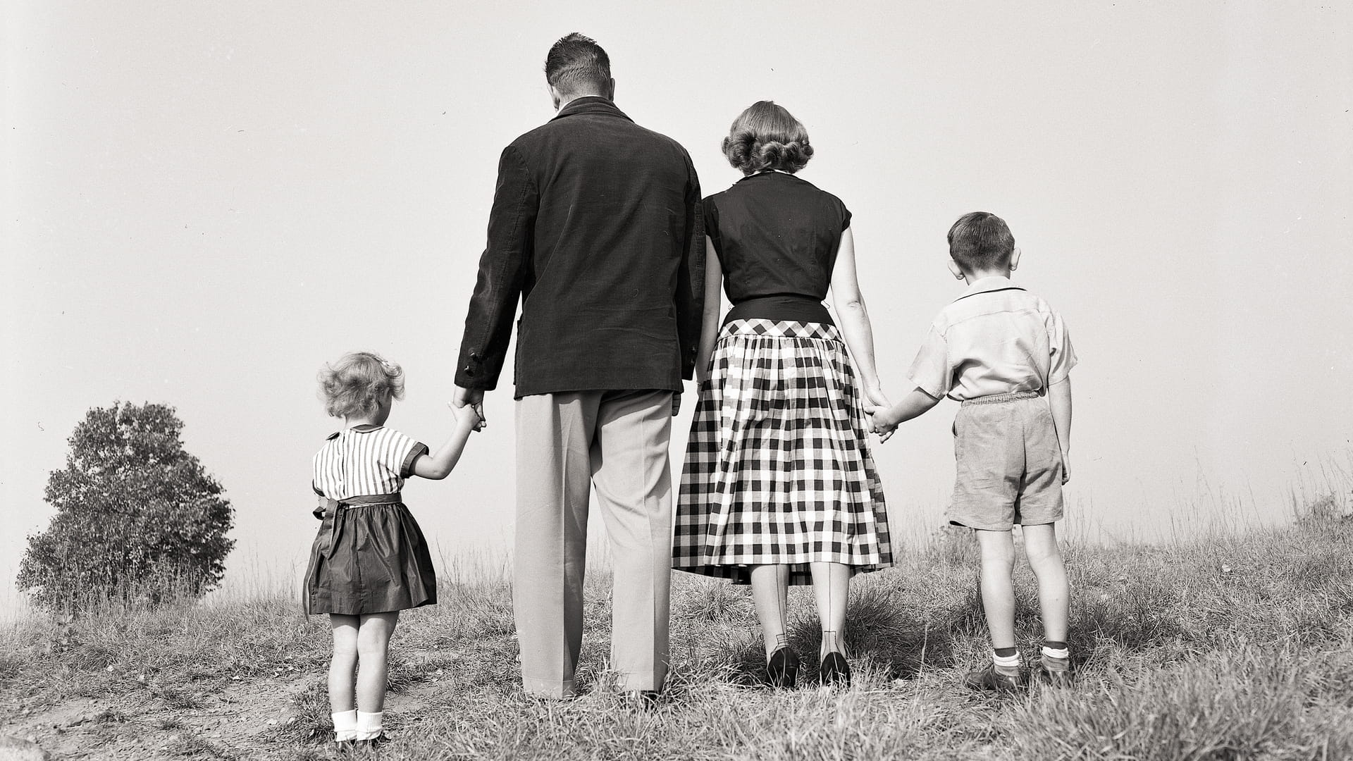 Een foto uit de jaren 50 van twee volwassenen en twee kinderen die elkaars handen vasthouden. Je ziet enkel de achterkant van alle mensen in het beeld. 