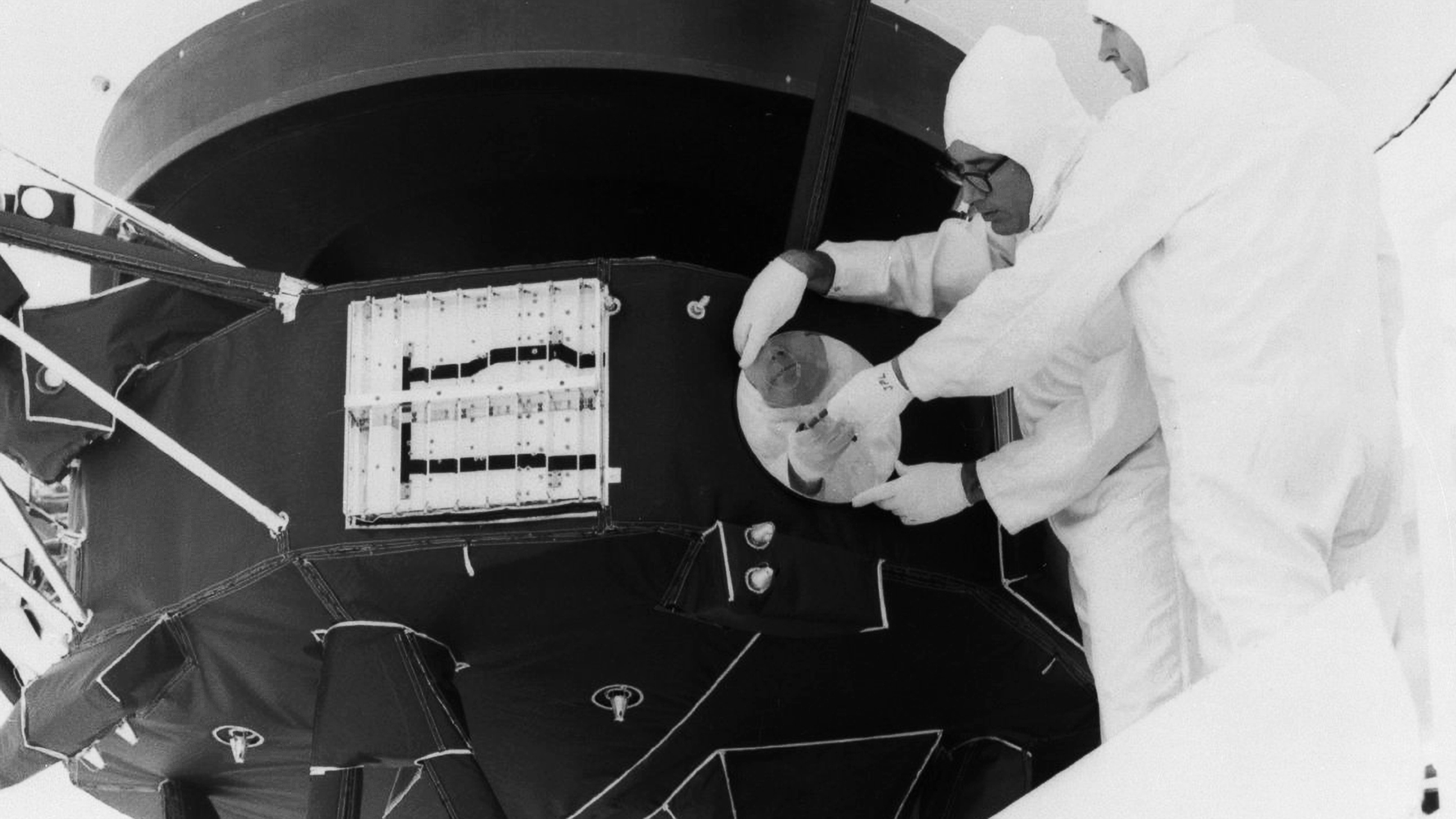 Zwart wit foto. 2 Technici van NASA gehuld in witte antistatische pakken bevestigen de gouden plaat aan de buitenkant van de ruimtesonde Voyager 2