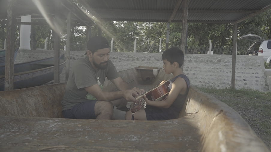 Carlos Choc en zijn zoontje zitten in een vissersboot, die aan wal ligt. Carlos leert zijn zoontje gitaar spelen.