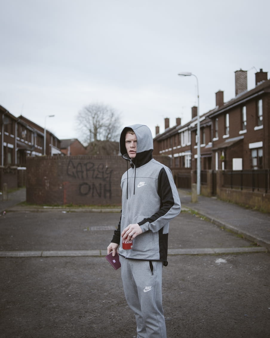 Een jongen gekleed in grijs joggingpak poseert met een Iers paspoort dat hij op straat heeft gevonden in Belfast. In zijn andere hand een blikje Cola.