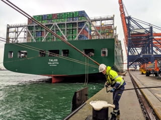 Een van de 'roeiers' legt een tros om een meerpaal. Links in beeld is de groene achtersteven van het containerschip Talos te zien. Rechts in beeld staan de kranen klaar om de containers van het schip te lossen