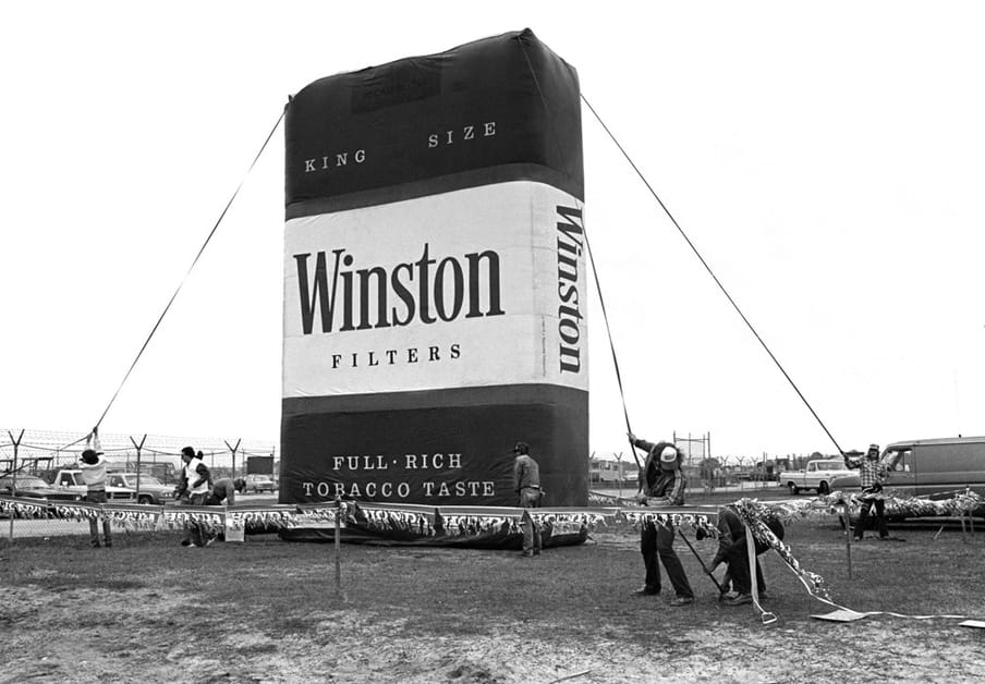 Werklui zetten een opblaasbaar Winston-sigarettenpak op voorafgaand aan de Daytona 500 in Florida, 1980. R.J. Reynolds Tobacco Company was in de jaren 80, voordat sigarettenreclame in Amerika werd verboden, de grootste sponsor van NASCAR. Foto: Robert Alexander / Archive Photos / Getty Images