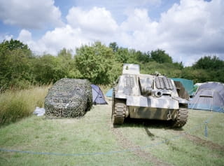 Auto met camouflage net erover, een neptank en op de achtergrond tentjes