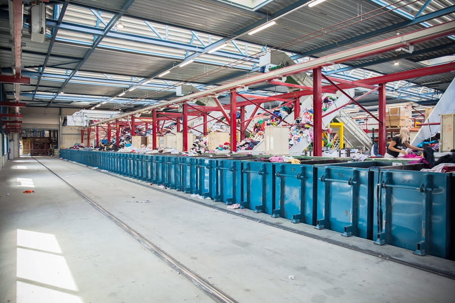 Een foto van een grote loods met een rij blauwe containers, hierachter zijn lopende banden met kleding te zien. 